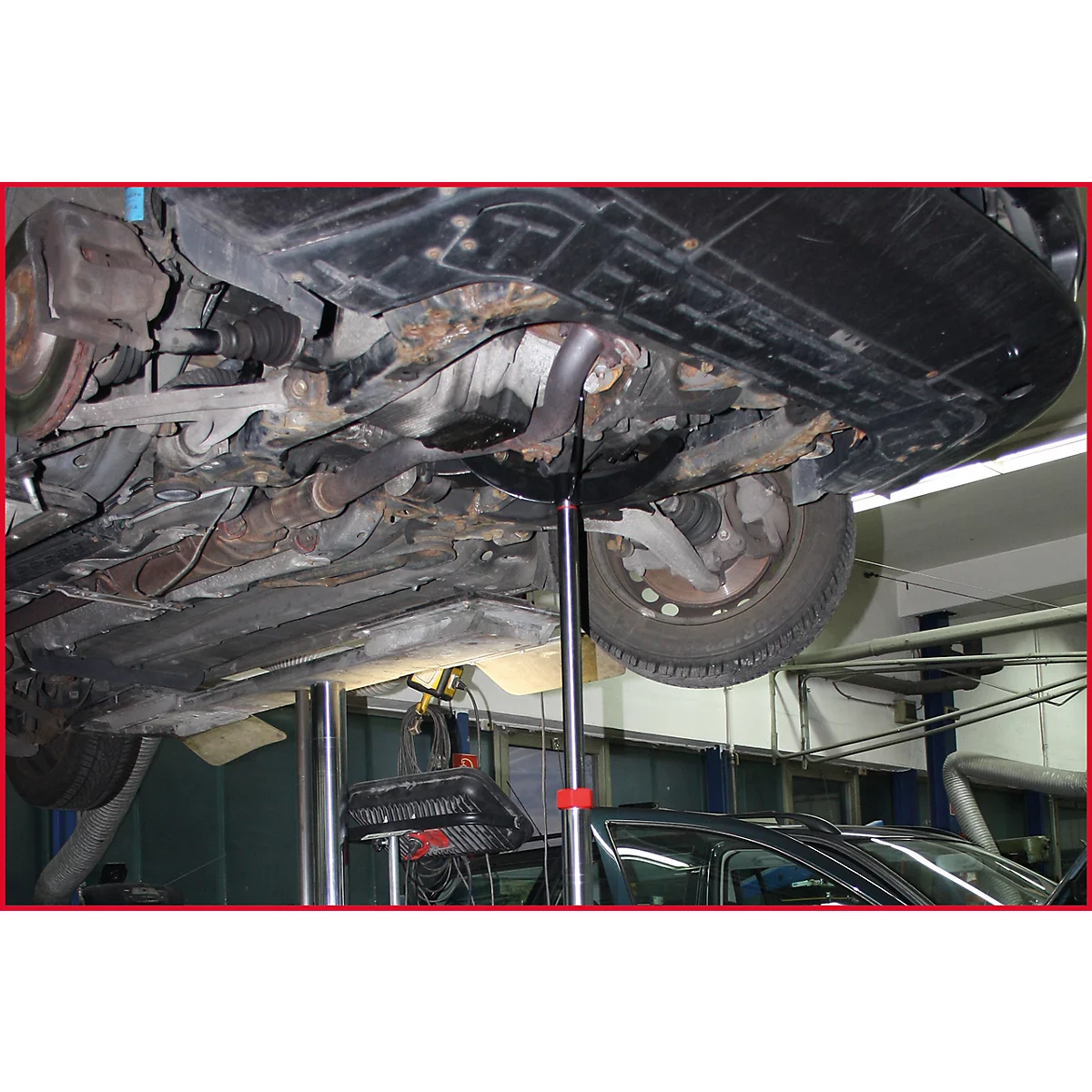 Blick von unten auf ein Auto, das auf einer Hebebühne steht. Der Unterboden, die Auspuffanlage und das Rad sind gut sichtbar.